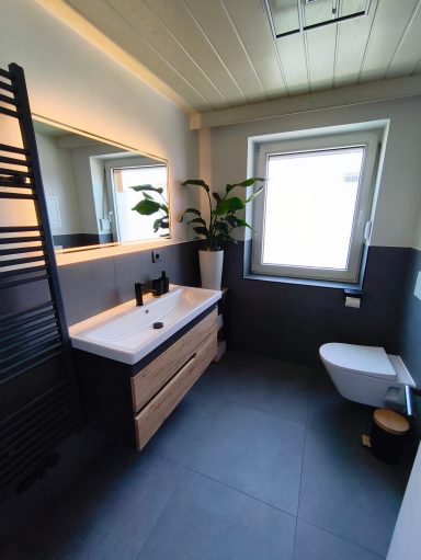 Helles, modernes Badezimmer mit großem beleuchtetem Spiegel, Holz-Waschtisch und Fenster in der Ferienwohnung Burgperle in Burghausen.